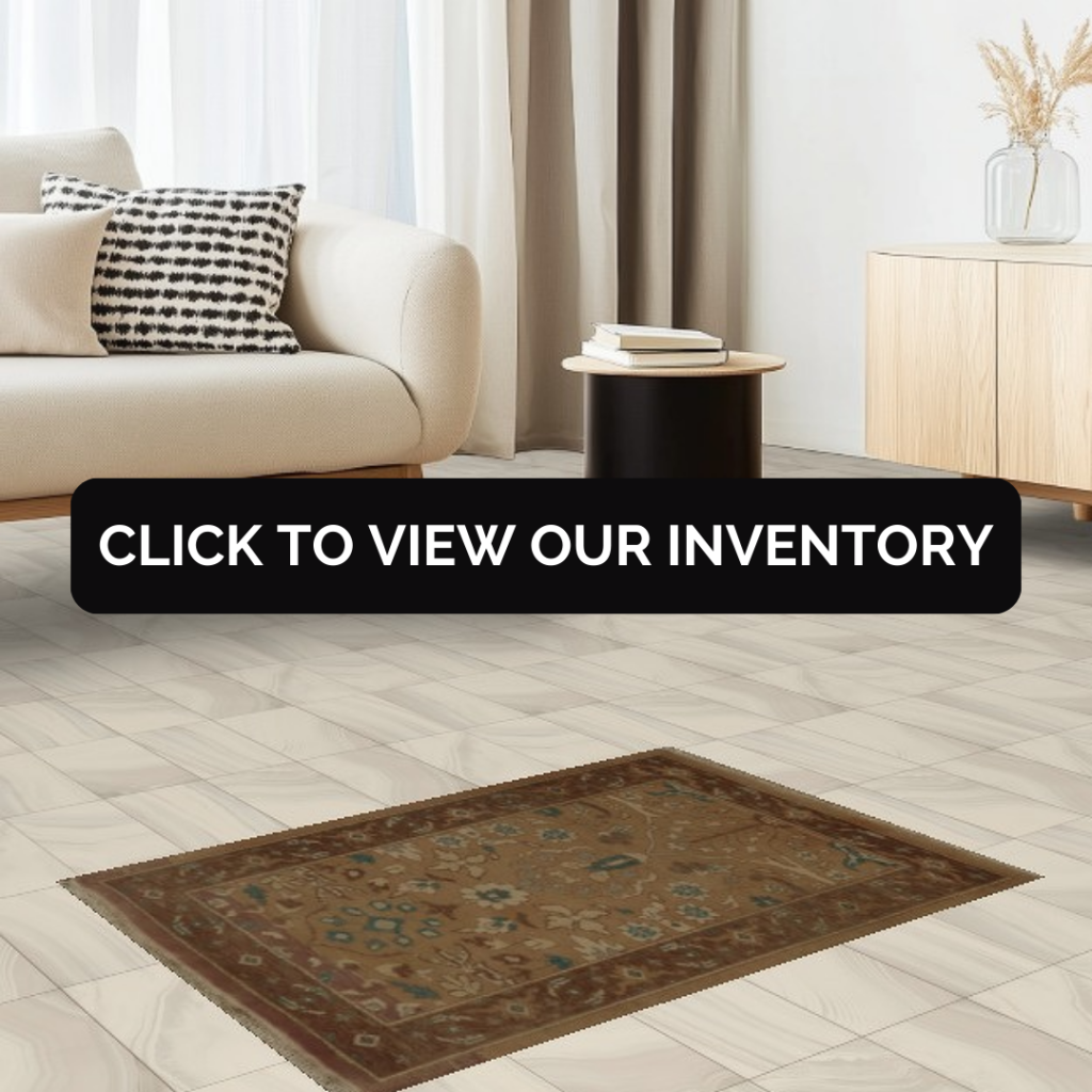 A light, modern living room with a cream sofa and a black-and-white patterned pillow sits beside a small round side table and a light wood cabinet with a vase of dried stems. A brown traditional-style area rug with intricate floral motifs rests on pale tile flooring. A black banner across the center reads, “CLICK TO VIEW OUR INVENTORY.”