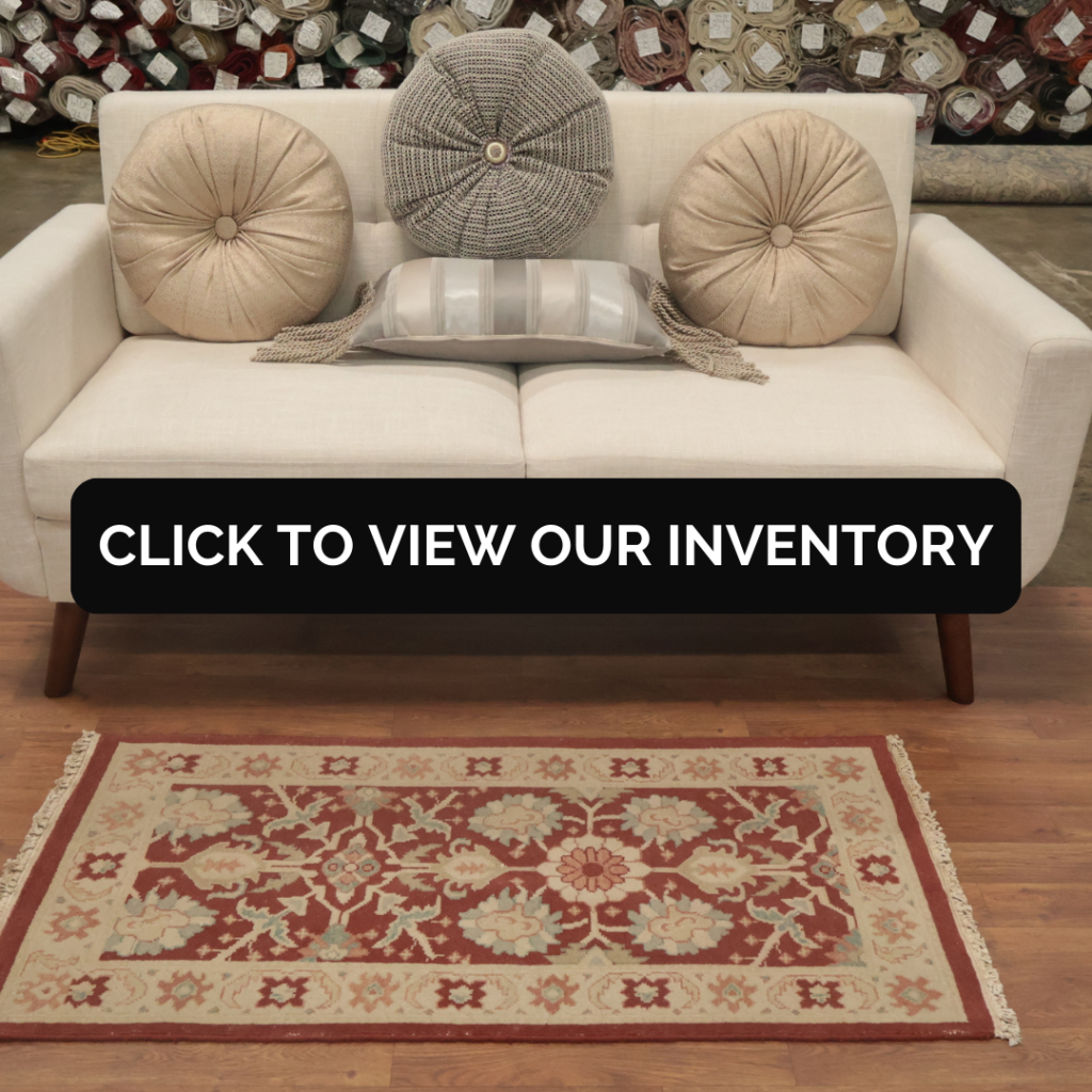 A cream-colored sofa with three round decorative pillows—two beige and one gray—sits on a hardwood floor in front of stacked fabric rolls. A red and beige floral-patterned area rug with a bordered design lies centered beneath the sofa. A black banner across the middle reads, “CLICK TO VIEW OUR INVENTORY.”