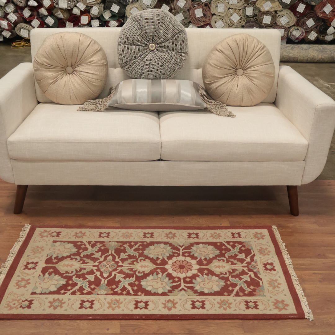 A cream sofa with three textured round pillows is positioned above a red and beige floral area rug on a hardwood floor. Fabric rolls are stacked along the back wall, suggesting a showroom setting. The rug features a detailed border and central floral medallion design.