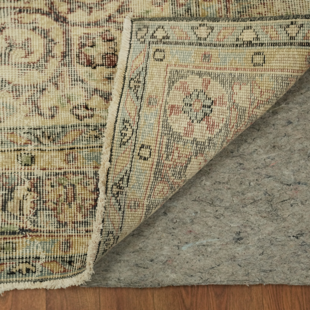 A close-up of a rug corner folded back shows the detailed backing and stitched border with a gray rug pad underneath. The intricate pattern and fringe edge are visible along the fold.