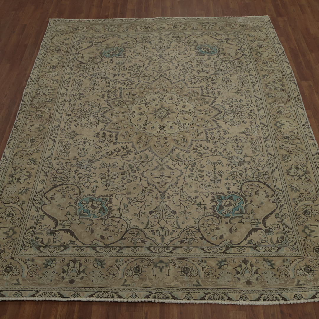 A beige vintage-style rug with a large central medallion and delicate floral patterns is spread across a hardwood floor. Subtle teal accents appear within the ornate design.