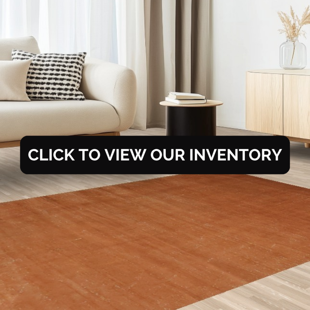 A modern living room with a cream sofa, black side table, and light wood cabinet, showcasing a solid rust-colored rug with subtle texture. A black banner across the center reads “Click to view our inventory.”
