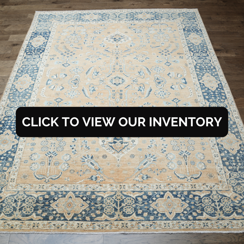 A large traditional-style rug in beige and soft blue tones with intricate floral patterns and a detailed border, laid on a hardwood floor. A black banner across the center reads “Click to view our inventory.”