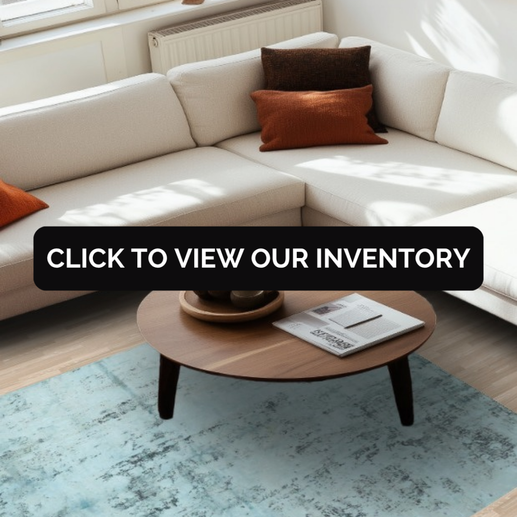 A bright living room with a white sectional sofa and wooden coffee table, showcasing a soft blue distressed-style rug. A black banner across the center reads “Click to view our inventory.”