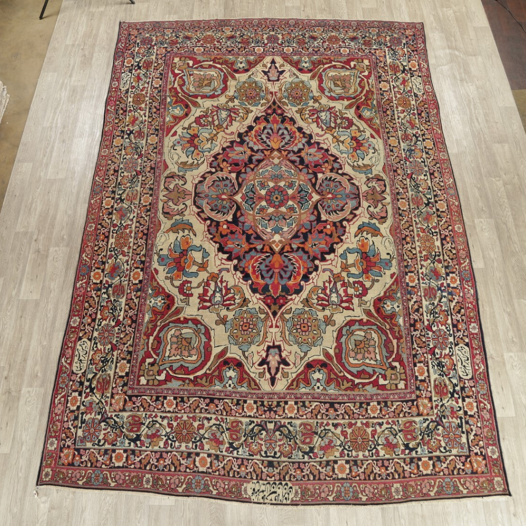 A traditional Persian-style rug with an ornate central medallion and floral motifs in red, cream, and navy tones, displayed on a light wood floor. The detailed border frames the symmetrical design with intricate patterns.