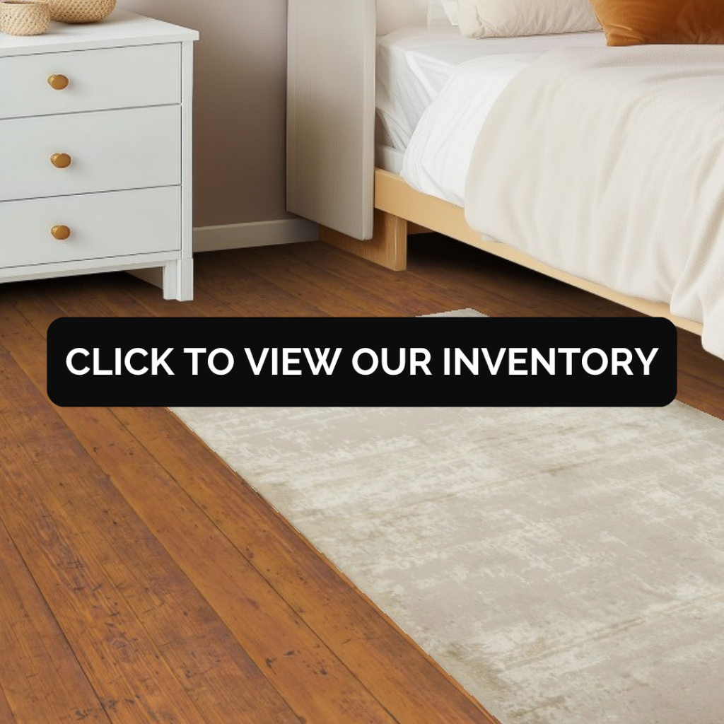 A bedroom scene with a wooden bed, white bedding, and a small nightstand, featuring a soft neutral rug in light beige tones. A black banner across the center reads “Click to view our inventory.”