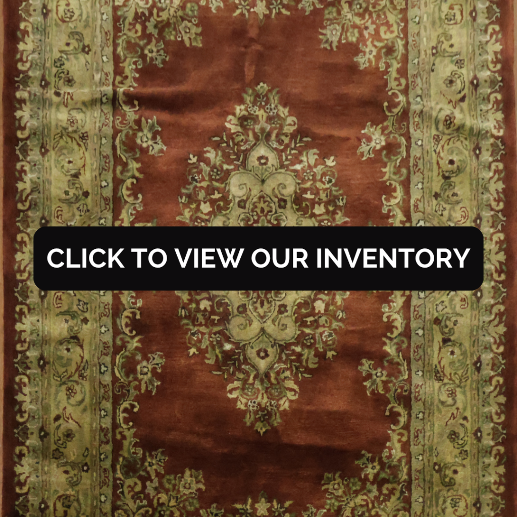 A traditional-style rug with a light green background and beige floral patterns, bordered with red and green accents, placed in a bright living room. A black banner across the center reads “Click to view our inventory.”