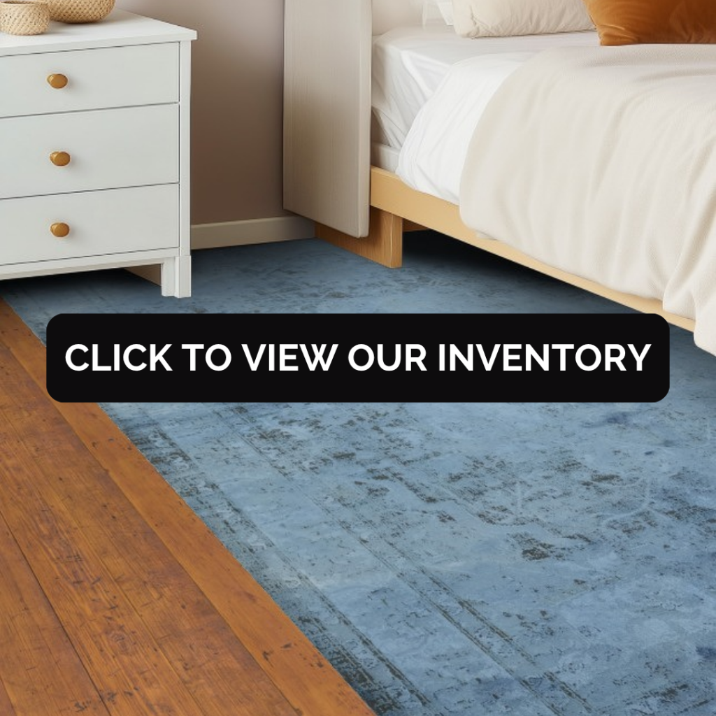 A bedroom scene with a wooden bed frame, white bedding, and a nightstand, featuring a muted blue rug with a distressed, vintage-style texture. A black banner across the center reads “Click to view our inventory.”