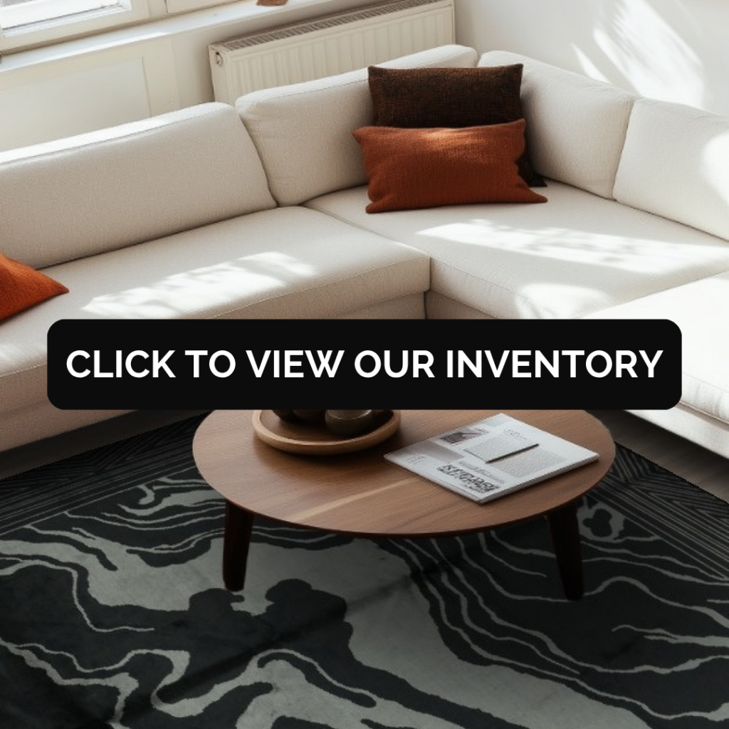 A cozy living space with a cream sectional sofa, warm-toned accent pillows, and a round wooden coffee table on a dark abstract rug. A centered black banner displays the text “Click to view our inventory.”